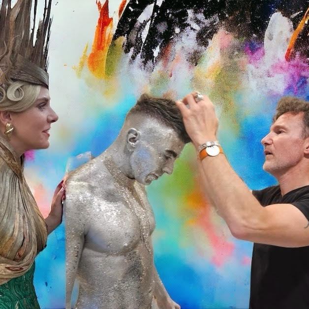 Cedric styling the hair of a male who's body is completely spray painted in silver. A light skinned woman in a spiky headdress looks on.