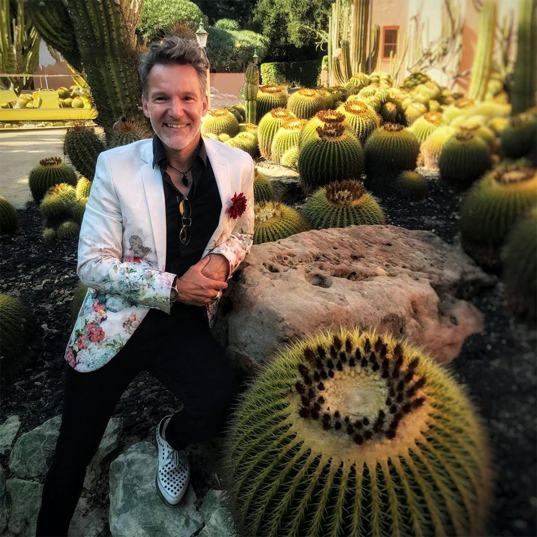 A light skinned man with wavy hair dressed in a white blazer and black slacks posing inside a garden of cactus.
