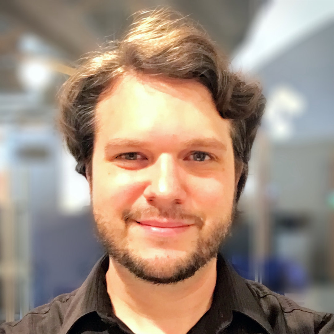 A smiling, youthful looking man with wavy brown hair and a slight beard.