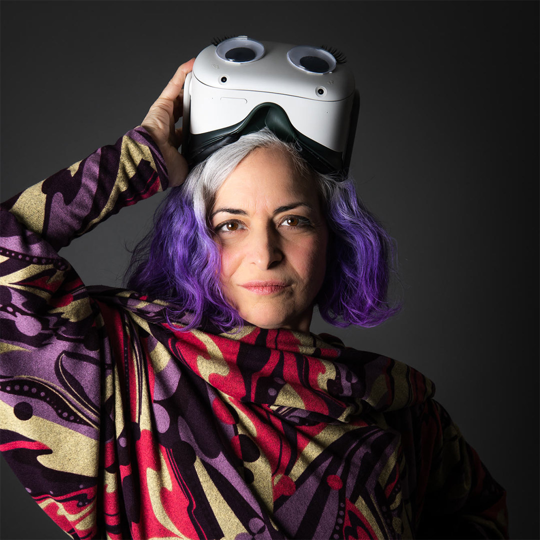 A middle aged woman with vibrant purple hair, wearing a colorful dress, holding a VR headset with large googly-eyes affixed to the front of it.