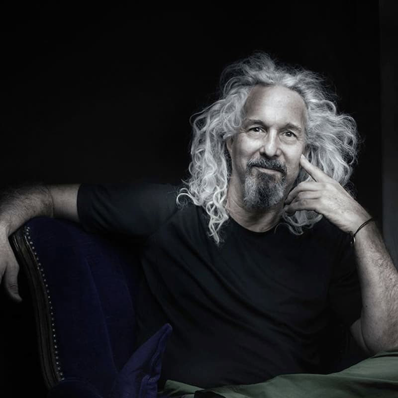 An older man with long gray hair and salt and pepper beard, wearing black T shirt and subtly smiling.