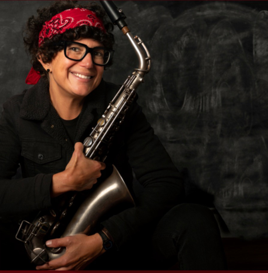 Woman with olive skin and wearing a red bandana in her curly black hair. She has on thick rimmed eyeglasses and is holding a saxophone.